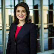 A person in formal attire, wearing a navy blazer and red top, smiles in front of large windows with a cityscape view.