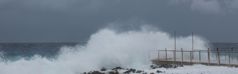 Waves crashing against rocky shore with a metal railing, under an overcast sky, creating a dramatic seaside scene.
