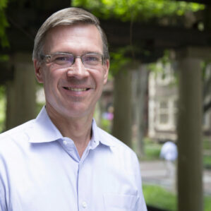 Witold Henisz smiling in a light colored button down outside in front of trees and brown brick buildings.