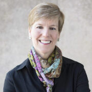 Katherine Klein Esg Headshot of a person with short hair, wearing a colorful scarf and a dark top, smiling against a neutral background.