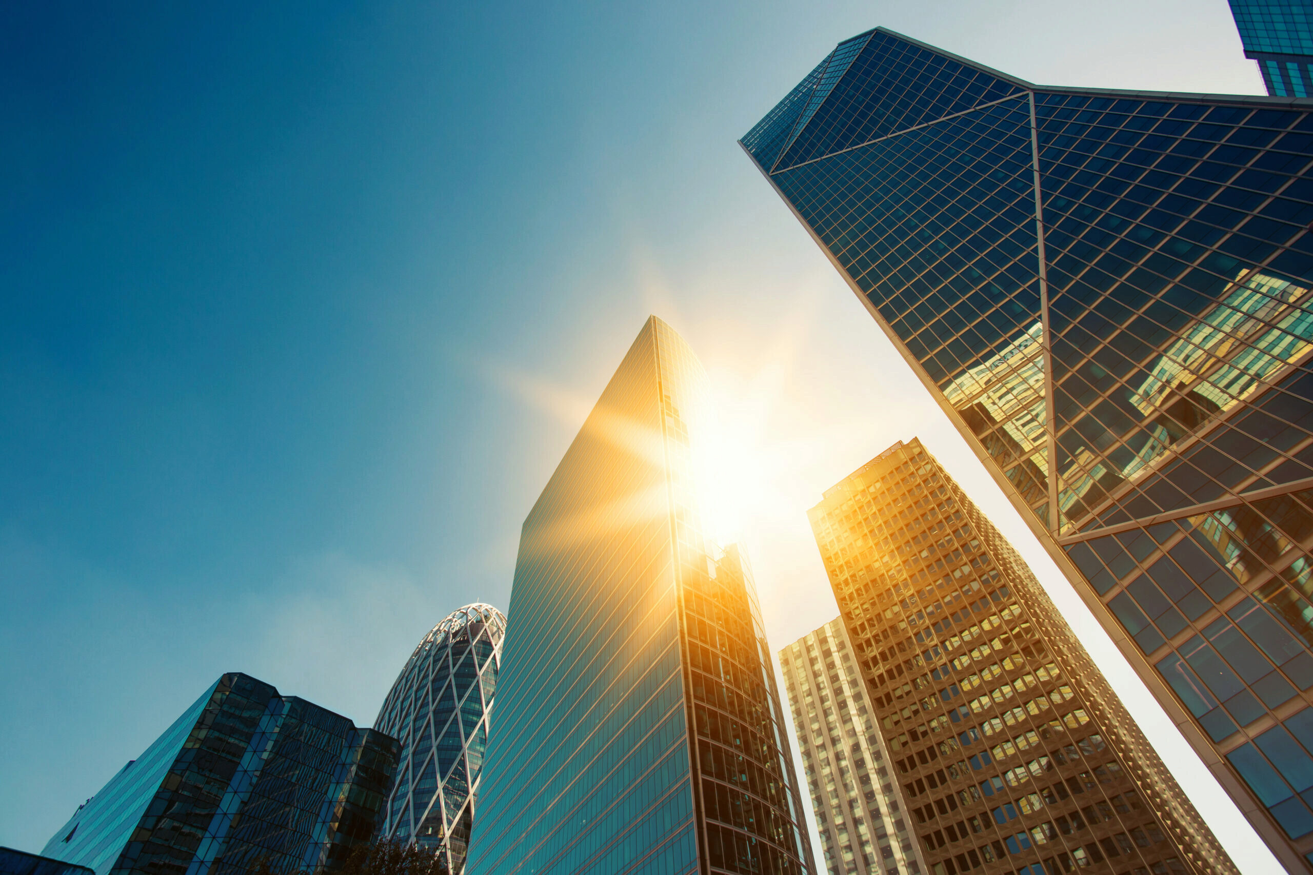 A cluster of modern skyscrapers with reflective glass facades, with sunlight shining brightly between two buildings against a clear blue sky.