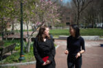 Two people walking and talking along a brick pathway in a park area, with pink flowering trees and green grass in the background.