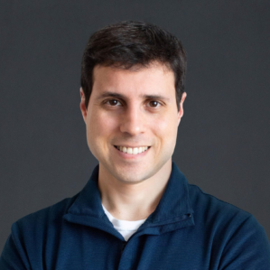 Headshot of a person smiling, wearing a dark blue shirt, against a plain dark background.