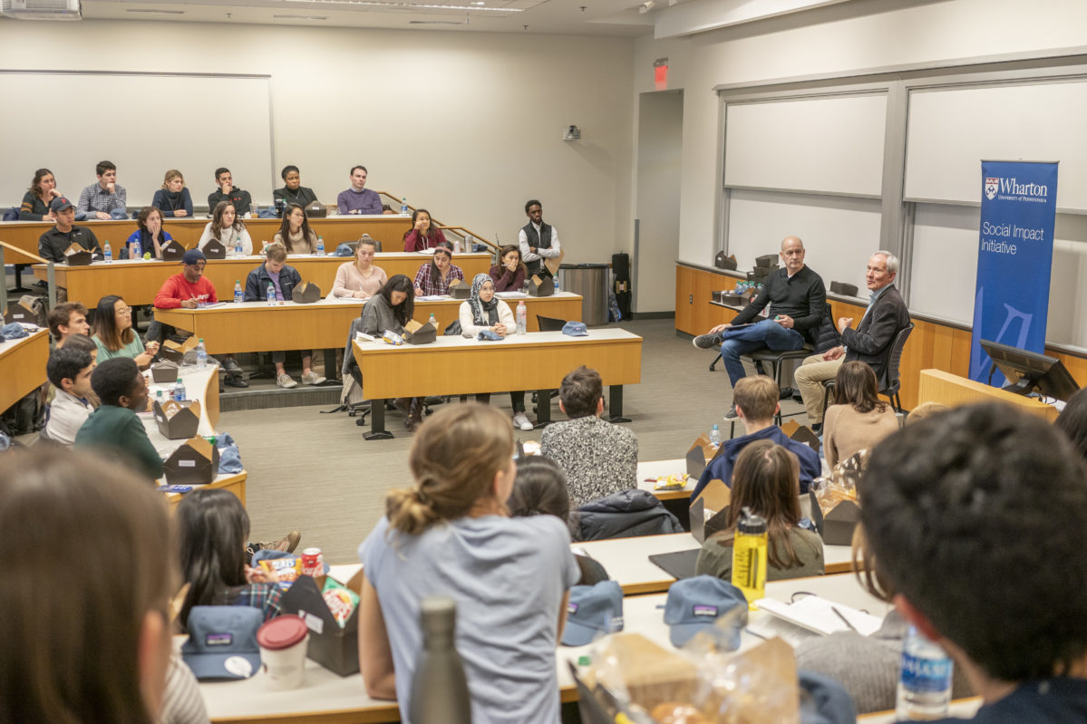 A classroom setting with a group of students seated in tiered rows, listening to two speakers sitting at the front. A banner reads "Wharton Social Impact Initiative."