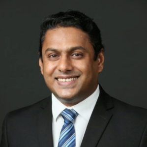 A person in a business suit and striped tie smiling, set against a dark background. Headshot.