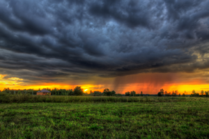 Sunset Storm A dramatic landscape showing a green field under a stormy sky with dark clouds and a distant sunset. A house is visible near a line of trees, with rain falling in the background.