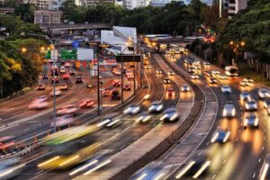 Untitled Design 1 A busy multi-lane highway with blurred vehicle lights, indicating fast-moving traffic, surrounded by greenery and urban buildings.