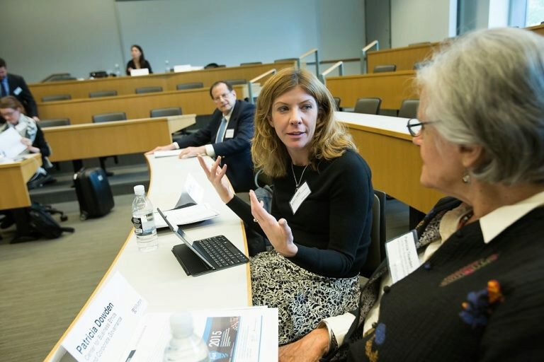A group of people in a conference or academic setting, seated around a table in a lecture hall. They are engaged in a discussion, with laptops and papers visible.