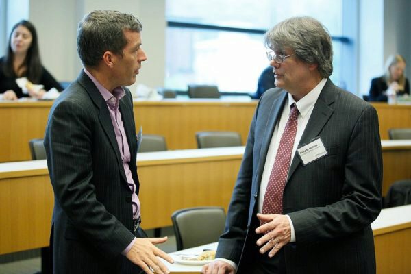 Two people in business attire are engaged in conversation in what appears to be a conference or meeting setting, with others in the background.