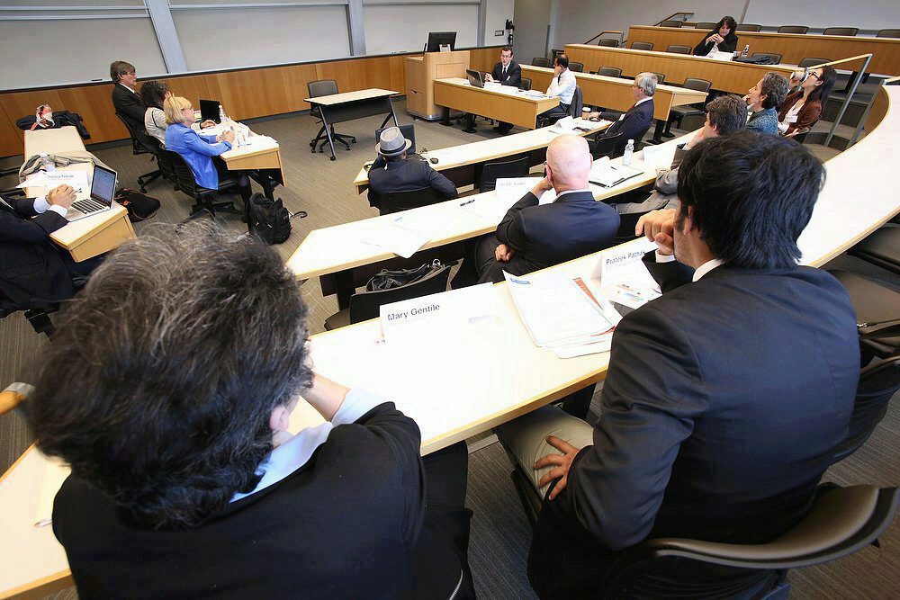 A group of people is seated in a tiered classroom or lecture hall, engaged in discussion. Participants use laptops and papers at their desks.