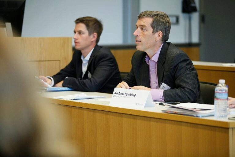 Two individuals in business attire are seated at a conference table with a nameplate showing "Andrew Spalding." They appear engaged in a discussion or presentation.