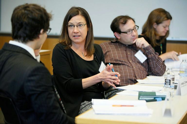 A group of people in a conference or meeting setting. One person is speaking while others listen and take notes. Papers and water bottles are on the table.
