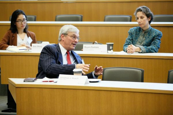 Three people in a lecture hall, seated at desks. The person in the foreground is speaking and gesturing, while the other two listen attentively. Name placards and coffee cups are visible.
