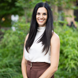 Ashley Sogge smiling in a white blouse and brown pants outside in front of foliage.