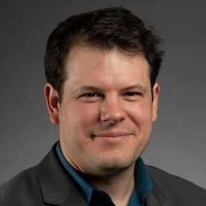 Dan Petty smiling in a blue button down and dark grey suit in front of a grey background.