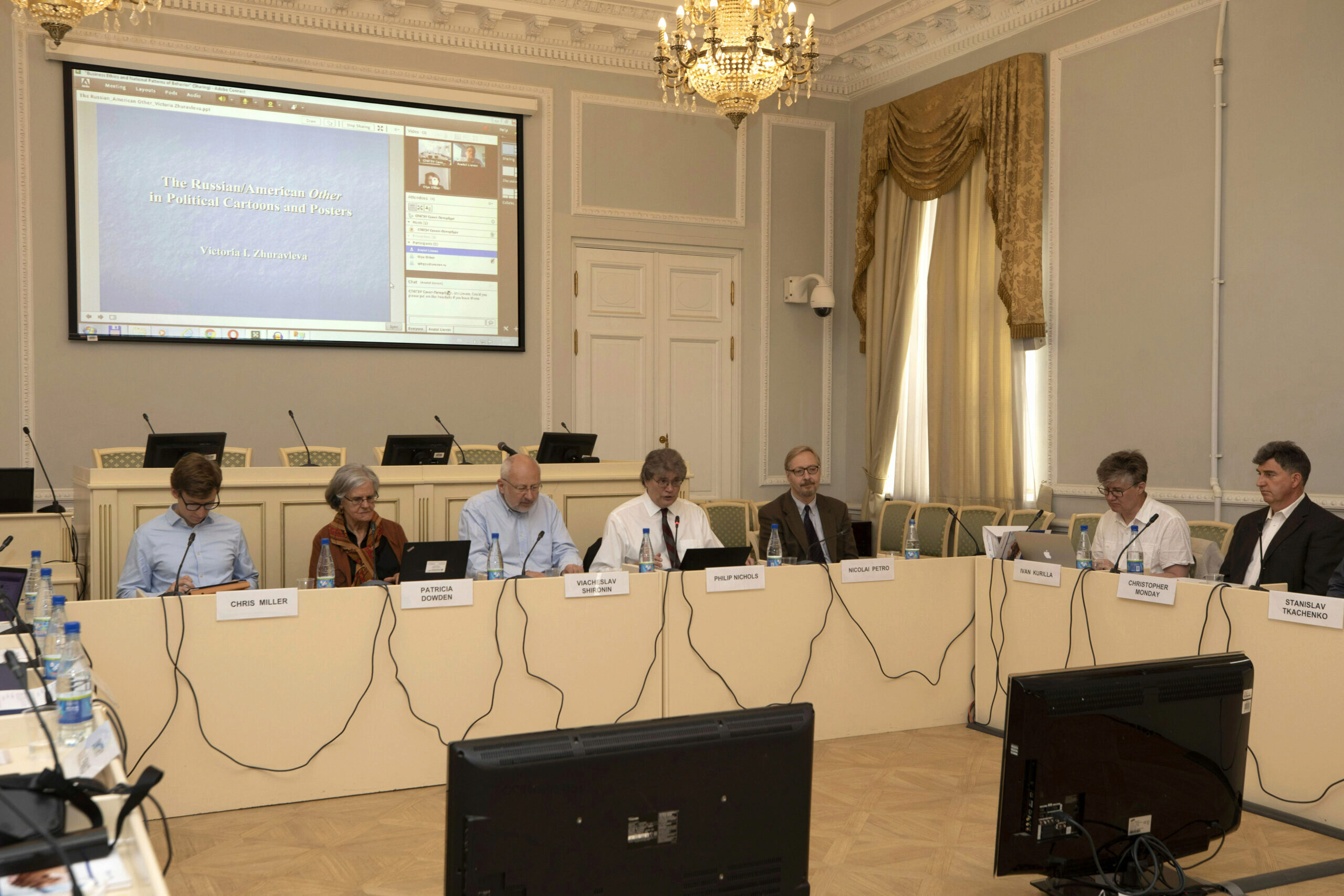A conference room with seven people seated at a long table, each with nameplates, microphones, and laptops. A large screen displays a presentation titled "The Russian/American Other in Political Cartoons and Posters." The room is elegantly decorated with chand