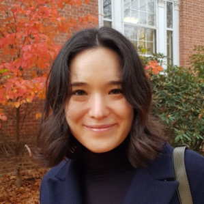 Elizabeth Saunders in a black turtleneck and blue jacket smiling outside in front of a brick building and a tree with orange leaves during the fall.