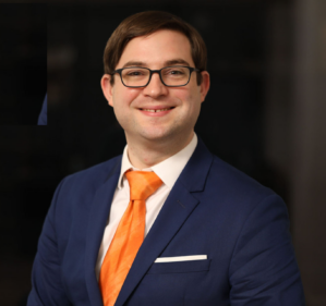 Evan Holownia Evan Holownia smiling in a white button down, orange tie, and navy blue suit against a dark background.