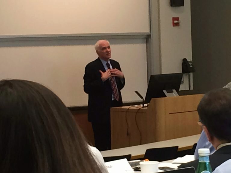 A person giving a lecture in a classroom setting, standing near a podium with a computer and microphone, gesturing while speaking to an audience.