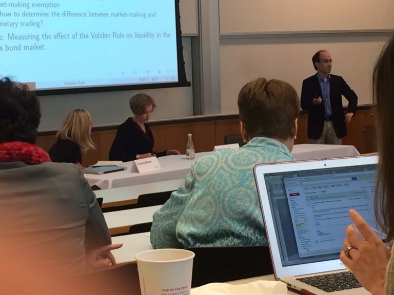 A person is presenting in a classroom or conference setting. Attendees sit at tables watching the speaker, with laptops and papers in view. A projector screen displays text.