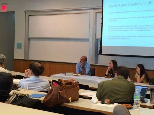 A panel of people sitting at a table in a classroom or conference setting, with an audience listening. A presentation slide is projected on a screen in the background.