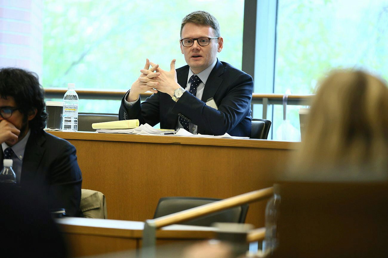 A person in a suit is seated at a desk, speaking and gesturing with their hands in a classroom or conference setting. A notebook and water bottle are on the desk. Other people are partially visible in the foreground.