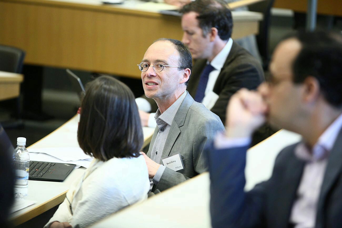 A group of people seated in a conference or seminar room, engaged in discussion or listening to a presentation. One person is facing the camera, wearing glasses and a name tag.
