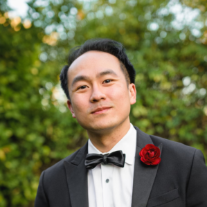 Michael Wong Michael Wong smiling in a black tuxedo, white button down, black bowtie, and red flower outside in front of foliage.