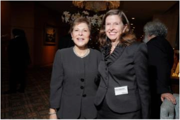 Two people in formal attire smiling at what appears to be an indoor event, with a chandelier and floral arrangement in the background.