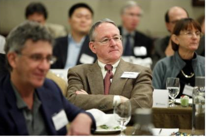 A group of people seated at a conference or meeting, with one person in focus sitting at a table, wearing a suit and glasses, listening attentively.