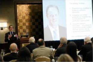 A person giving a presentation in a conference room, with an audience seated. A projection screen displays a slide with a person''s image and text.