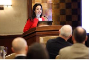 A person in a red top speaking at a podium with a microphone, addressing an audience in a conference or seminar setting.