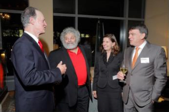 Four individuals in formal attire are engaged in conversation at a social event. One person is holding a glass of wine.