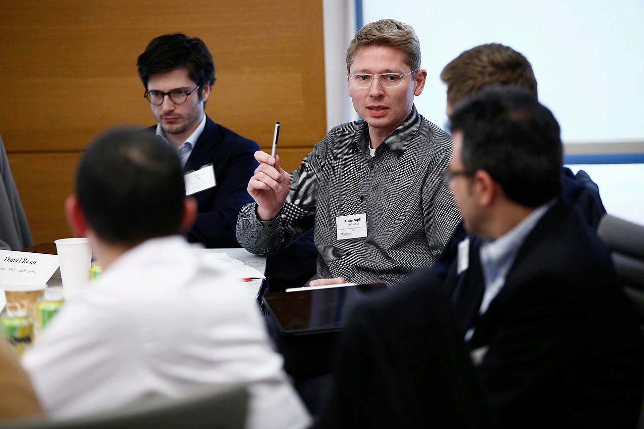 A group of people seated around a table during a meeting or discussion. One person is speaking and gesturing with a pen. Name tags are visible on their clothing.