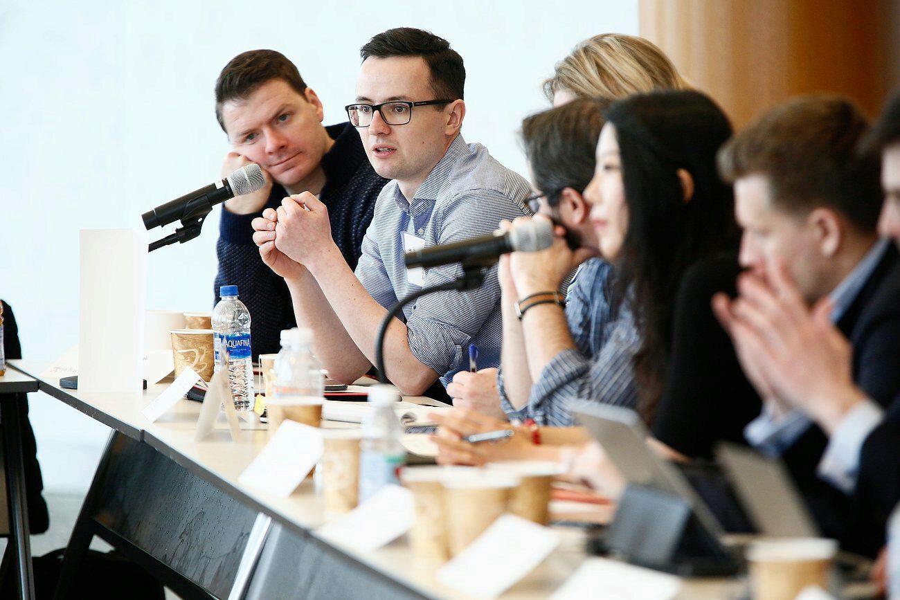 A group of people sitting at a long table engaged in discussion. They have microphones, notepads, and water bottles in front of them, indicating a panel or meeting setting.