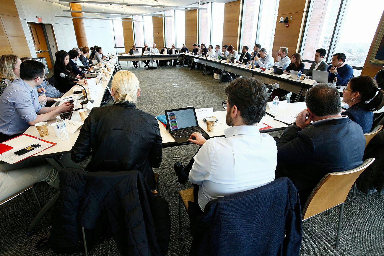A large group of people seated around a U-shaped conference table in a modern meeting room, using laptops and taking notes.