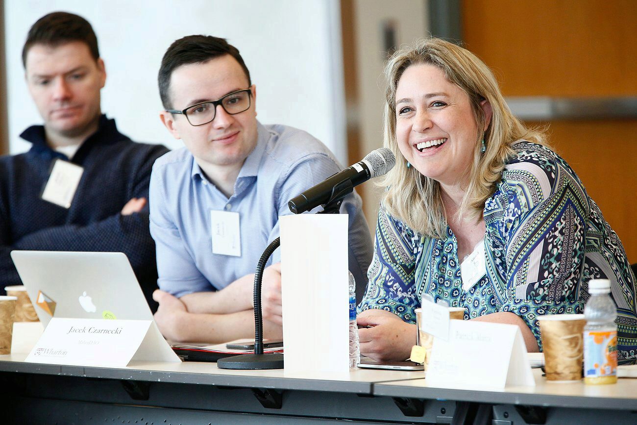 A group of people sitting at a conference table. One person is speaking into a microphone, smiling, with others listening attentively.