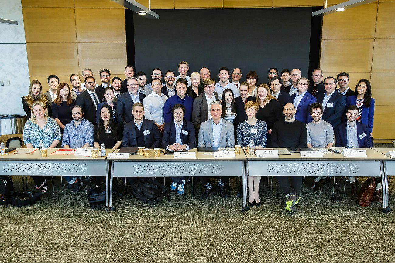 A group of people posing for a photo in a conference room, some seated at a table with nameplates and others standing behind them. The setting suggests a professional gathering or meeting.