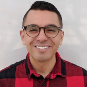 Richard Merino Richard Merino smiling in a red and black plaid button down against a cream wall.