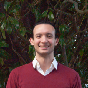 Shawn Yavari smiling in a red sweater over a white button down in front of a tree.