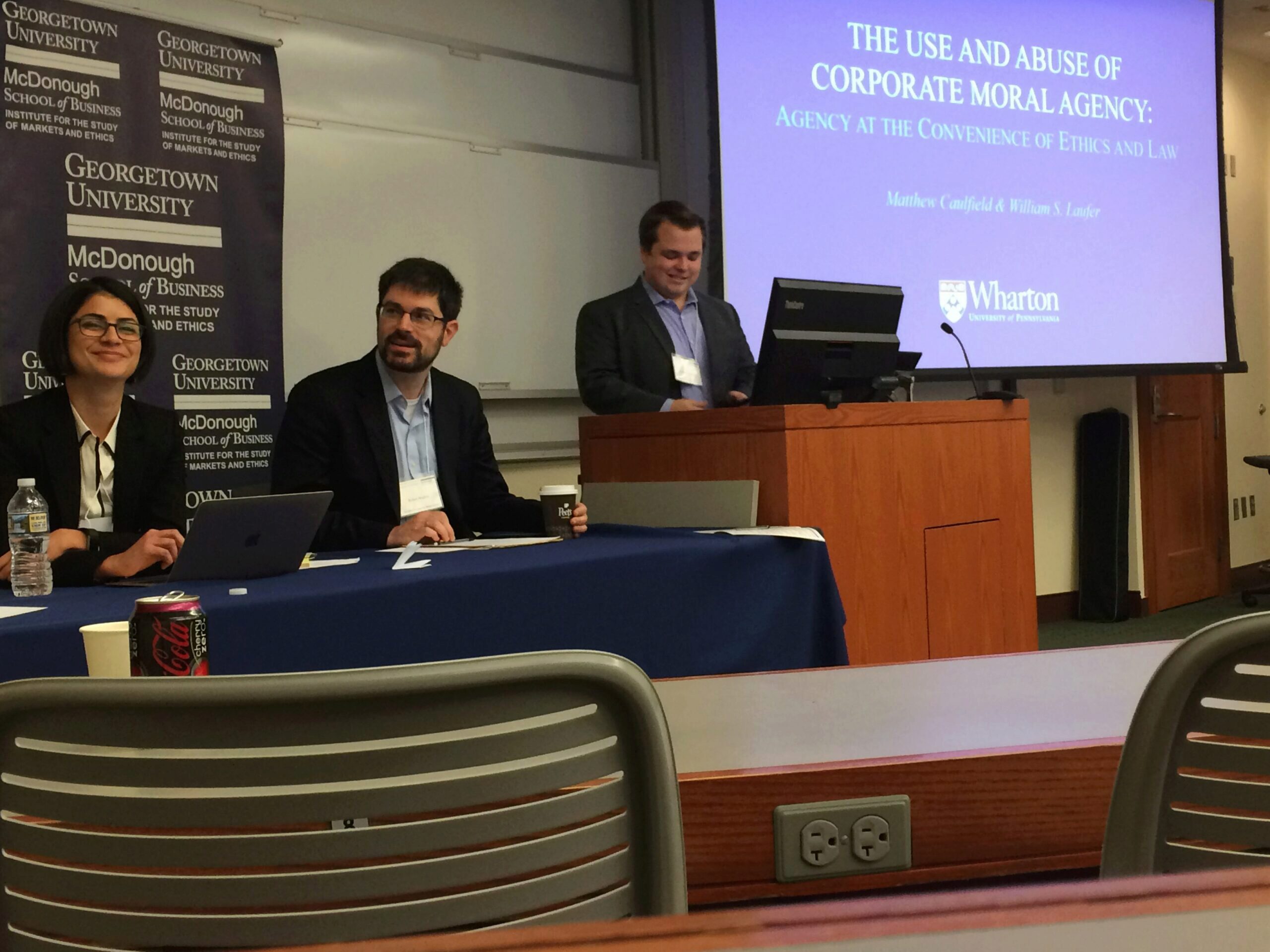 A panel discussion in a university classroom with three people. A projected slide titled "The Use and Abuse of Corporate Moral Agency" is visible.
