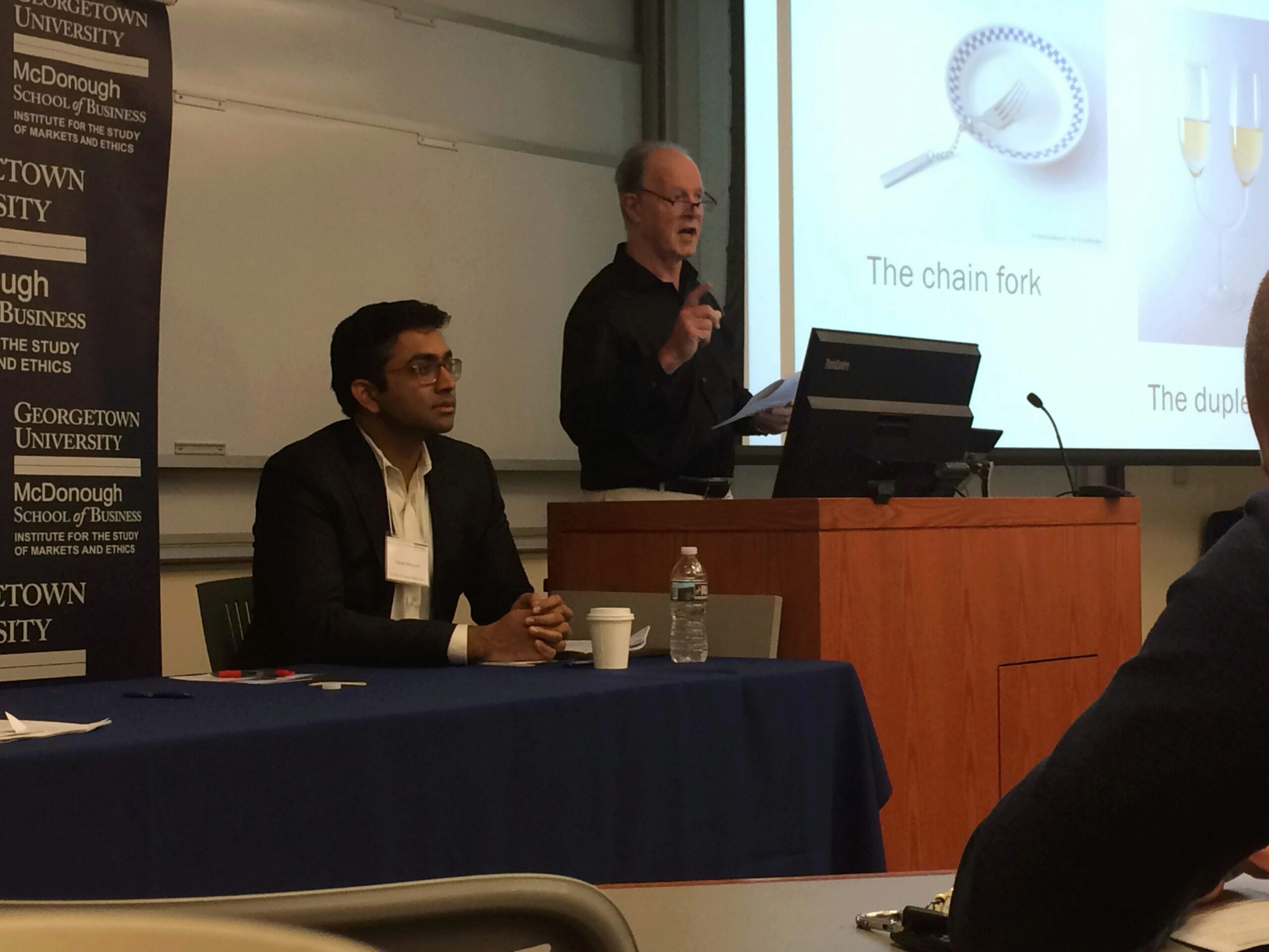 Two individuals in a Georgetown University McDonough School of Business setting; one is seated at a table and another is presenting at a podium. The projector screen displays images labeled "The chain fork" and "The duplex."