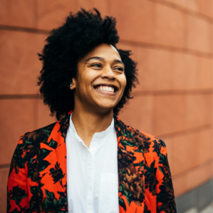 Trené Hawkins Tréne Hawkins smiling in a red and black floral blazer against a terra-cotta colored wall.