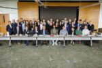 A group of people gathered for a formal meeting or event, posed for a group photo in a conference room. They''re dressed in business attire, seated and standing behind a long table.