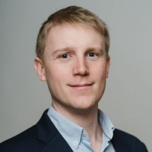 Professional headshot of man smiling in a navy blue suit jacket and baby blue button down in front of a grey backdrop