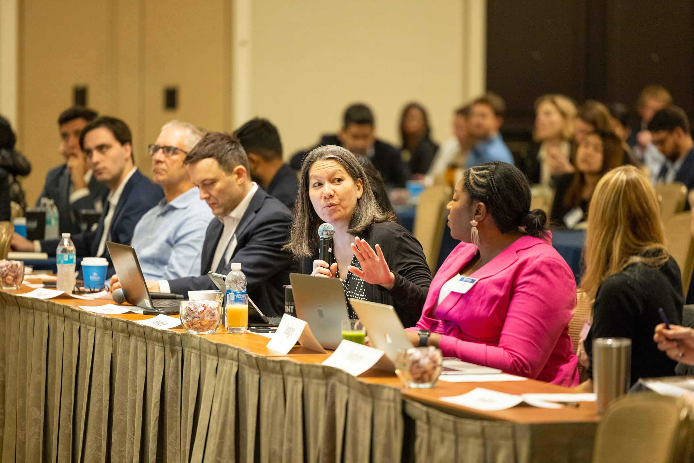 A person holding a microphone speaks at a conference or meeting. They are surrounded by others sitting at a table with laptops and documents, suggesting a discussion or presentation.