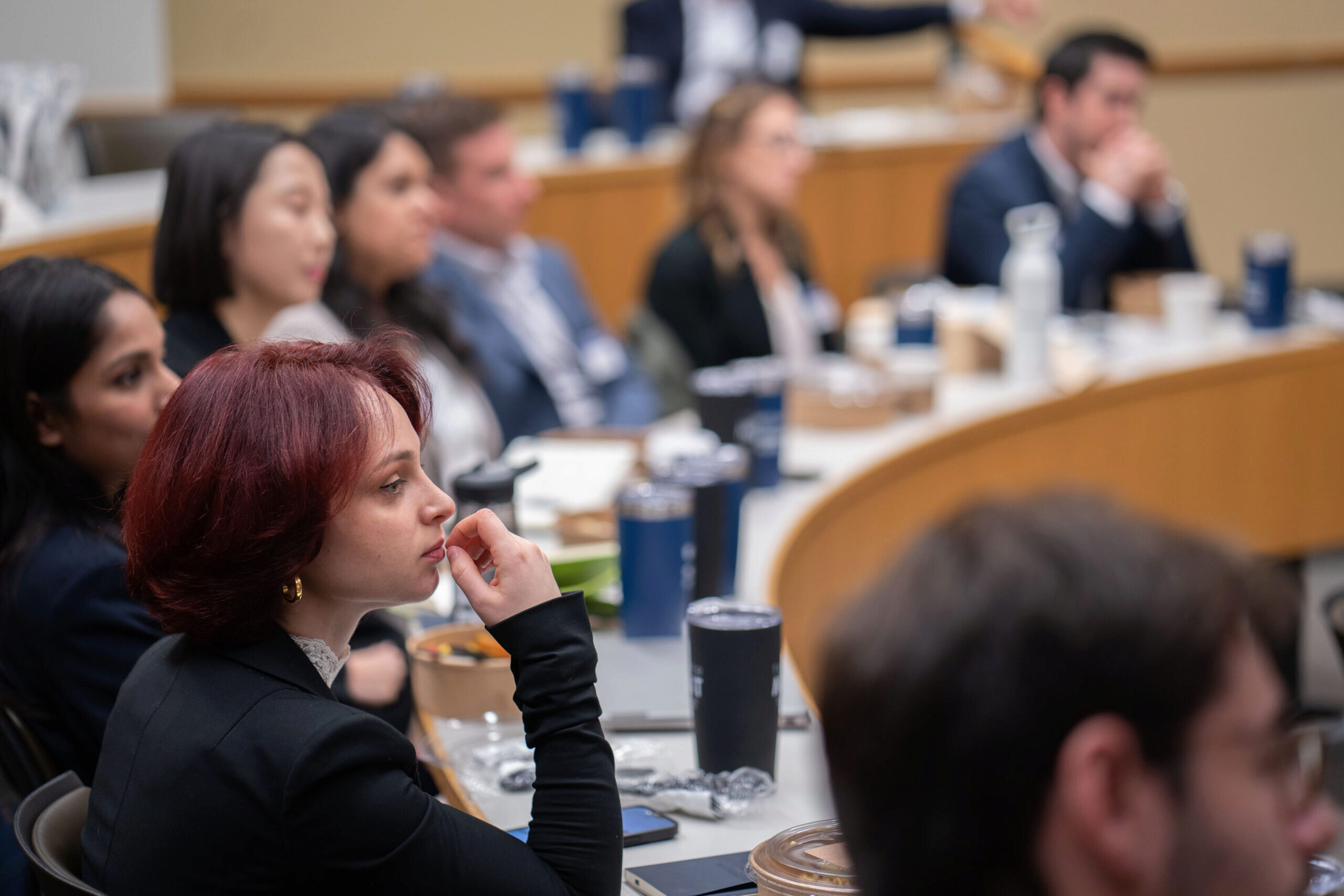 A group of people attentively listening in a classroom or lecture setting, seated in rows with desks, and various drinks on the tables.