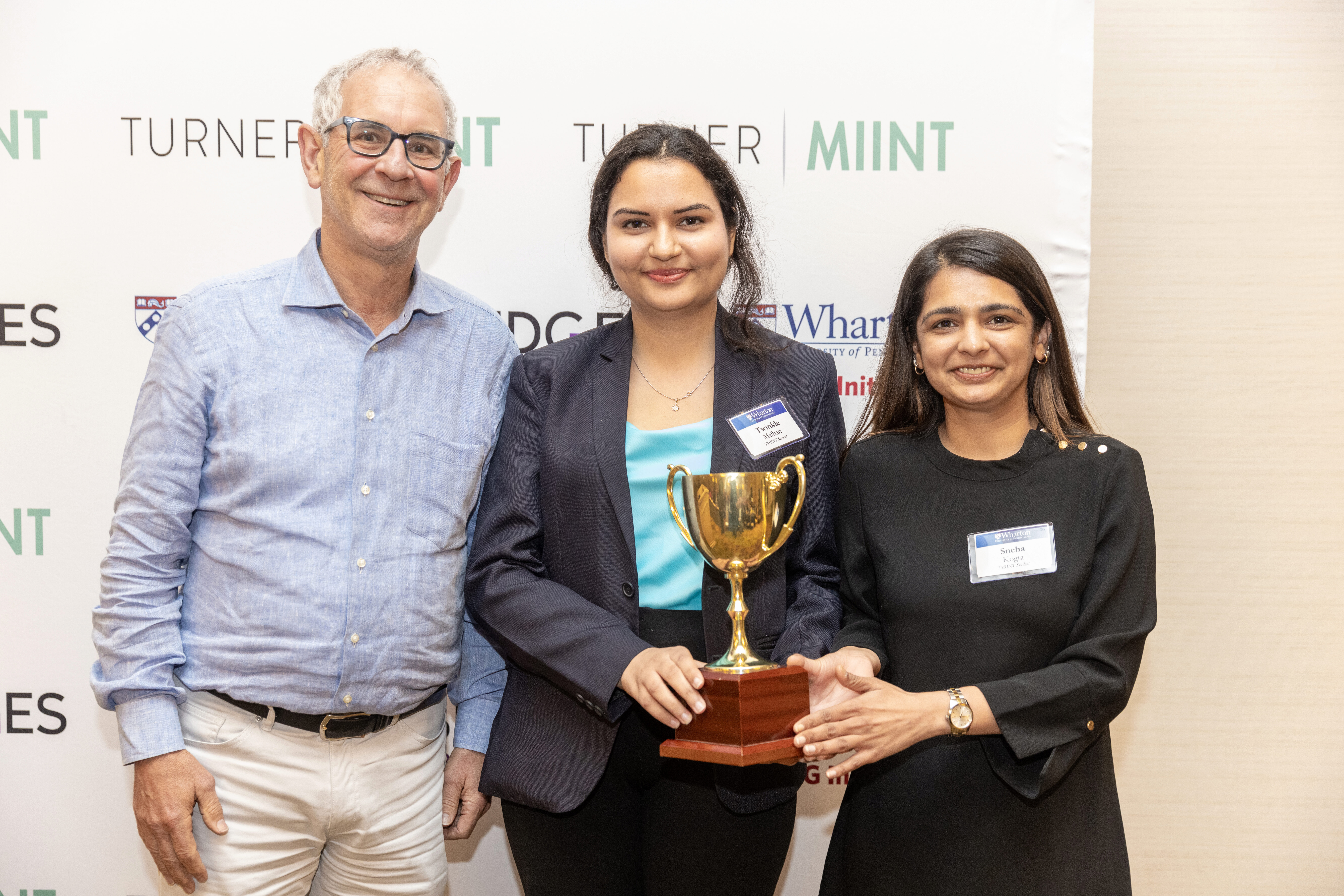 Saïd Business School Team 2023 TMIINT Ron Moelis poses with Twinkle Malhan and Sneha Kogta of the Saïd Business School Team. They're holding a gold tropy in front of a banner that says Turner MIINT, Wharton ESG Initiative, and Bridges Impact Foundation.