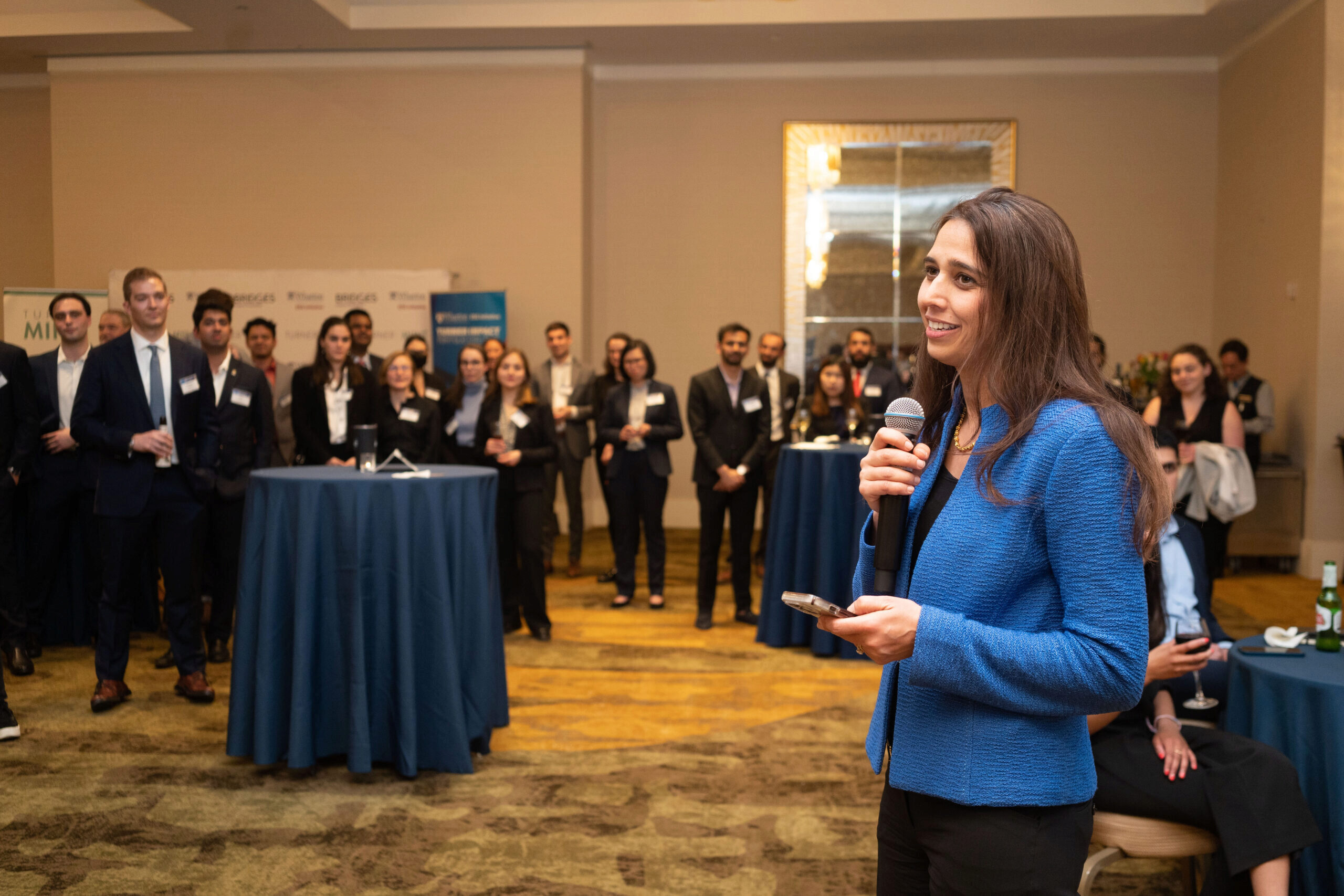 A person in a blue jacket holding a microphone speaks to an audience in a formal setting. The audience is standing, dressed in business attire.