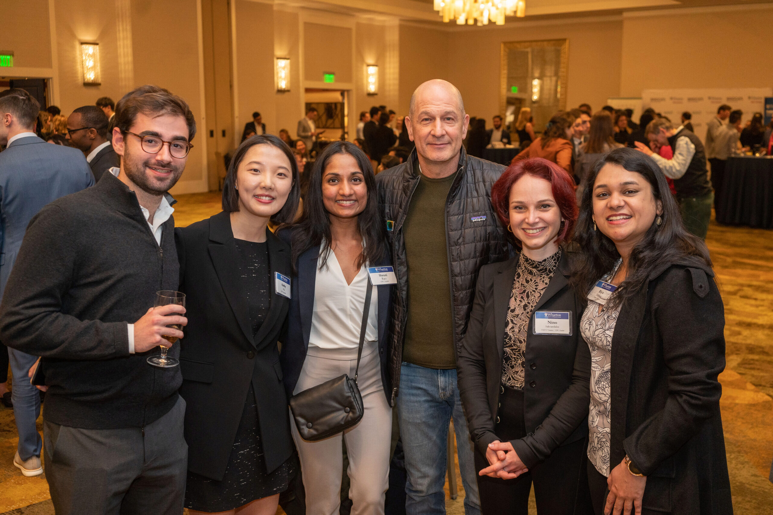 A group of six people posing together at an indoor event or networking gathering. They are well-dressed, with some wearing name tags, in a warmly lit room with other attendees in the background.